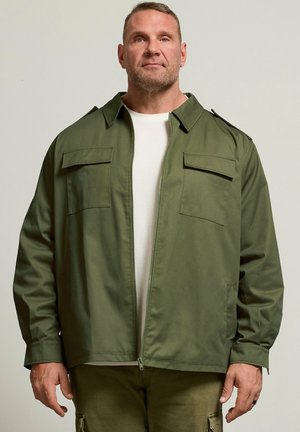 Mężczyzna w zielonej kurtce wojskowego stylu na białej koszuli, stojący na jednolitym jasnym tle, zwrócony przodem z neutralnym wyrazem twarzy.