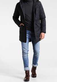 Homme portant un manteau à capuche noir, un col roulé sombre, un jean bleu clair retroussé et des bottes marron à lacets, marchant devant un fond blanc.