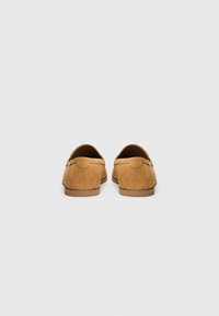 Mocasines de ante en tono marrón claro con punta redondeada, suela plana y detalles de costura en la parte posterior. Textura suave con un diseño minimalista.