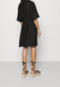 Donna con un vestito nero al ginocchio a maniche corte e sandali con zeppa neri a tracolla, in piedi davanti a uno sfondo semplice.