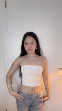 Jeune femme aux longs cheveux bruns portant un haut blanc sans bretelles et un jean bleu, debout contre un mur clair uni, les mains sur les hanches.