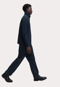 Camisa polo de manga larga en color navy con un cuello alto, combinada con jeans de mezclilla oscuros y botas negras de tobillo, mostrada en perfil lateral.