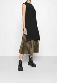 Svart ärmlös stickad klänning lager på lager över en veckad olivgrön kjol, i kombination med kraftiga svarta ankelstövlar och vita ankelsockor.