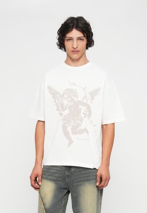 Een jonge man met krullend haar draagt een wit T-shirt met een beige cherub en de tekst "VERSPIJL CULTUUR NIET," in combinatie met vervaagde jeans.