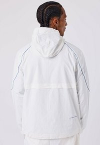 Witte hoodie-jack met blauwe accenten, lichte materiaal, zichtbare naden en een ontwerp van een achterkantzak, gezien vanuit de achterzijde.