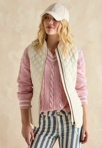 Elfenbeinfarbene, gestepfte Weste mit Reißverschluss, kombiniert mit einem rosa Kabelstrickpullover und hochtaillierten Jeans in Blau und Weiß mit Streifen.