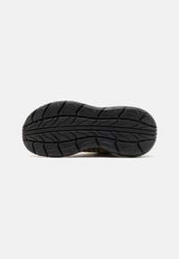Suela de zapato de goma negra texturizada con profundos surcos, que presenta patrones en zigzag y una marca en el lateral para mayor agarre y tracción.