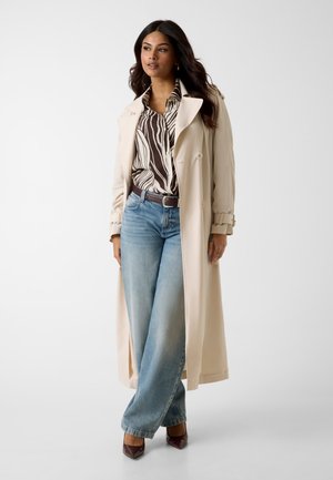 Femme portant un trench-coat beige sur une chemise à imprimé zèbre, un jean bleu à jambes larges et des escarpins bruns à bouts pointus, debout avec une jambe croisée.