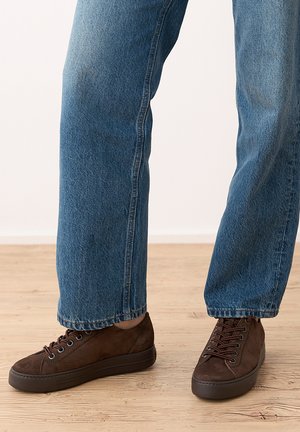 Baskets en daim marron foncé à semelles noires portées avec un jean bleu droit sur un sol en bois clair.