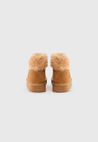 Par de botines de ante marrón con un borde de piel de peluche en la parte superior, vistos desde atrás sobre un fondo blanco liso.