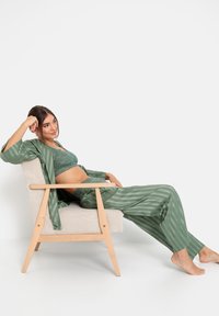 Groen gestreepte satijnen pyjama set met kanten accenten, bestaande uit een losse top en strakke broek, gecombineerd met een houten stoel op een witte achtergrond.