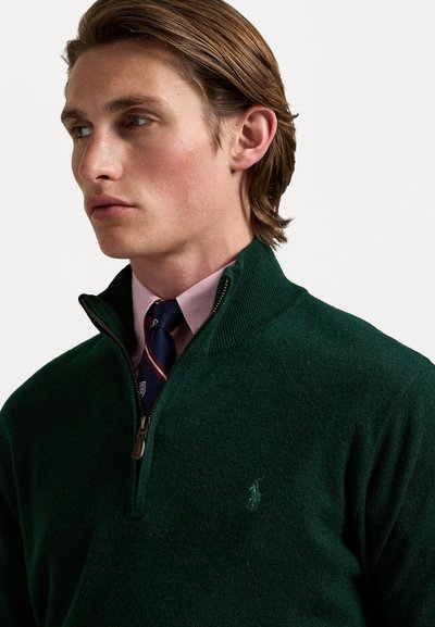 Sweater verde de lã com gola alta, meio zip e detalhes canelados. Usado sobre uma camisa rosa com uma gravata listrada em azul marinho. Detalhe sutil do logótipo visível.