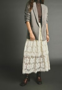 Blazer gris sur un col roulé marron, associé à une longue jupe en dentelle blanche avec des motifs floraux, et des chaussures marron à talons blocs.