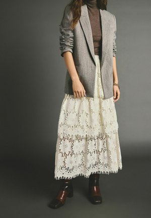 Blazer gris sobre un suéter de cuello alto marrón, combinado con una falda larga blanca de encaje con patrones florales y zapatos marrones de tacón grueso.