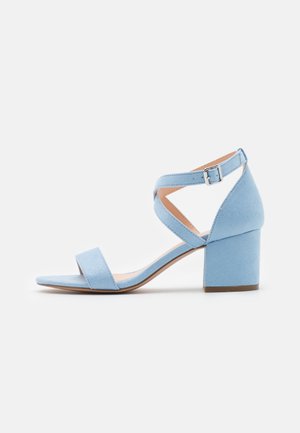 Sandale à talons blocs en daim bleu clair avec une sangle croisée à la cheville et une large bande à l'avant. Texture lisse et design ouvert au niveau des orteils.