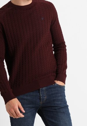 Mand iført en vinrød sweater med kabelstrik, ribstrikkede manchetter og kant, kombineret med mørkeblå jeans, med den ene hånd i lommen.