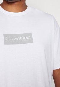 Camiseta de algodón blanca con un logotipo rectangular gris que lleva el texto "Calvin Klein" y "ESTABLECIDO EN 1968" en una fuente limpia. Cuello redondo clásico.