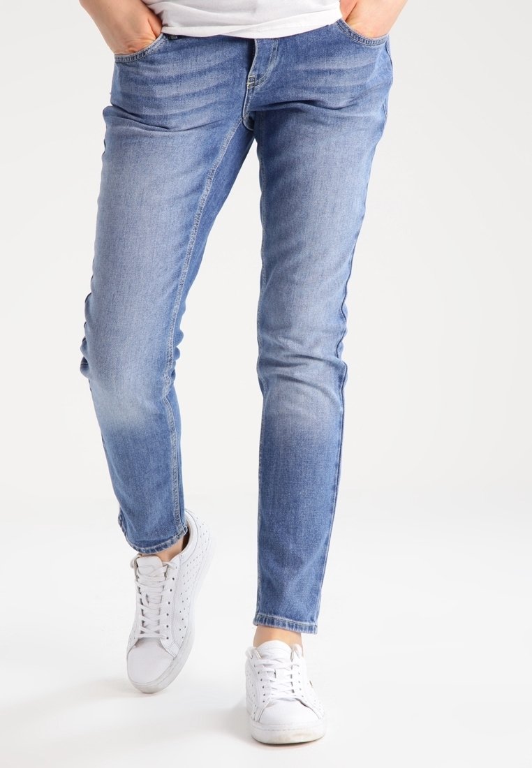 Osoba v světlomodrých slim fit džínách a bílých teniskách, s rukama v kapsách, stojící na bílém pozadí.