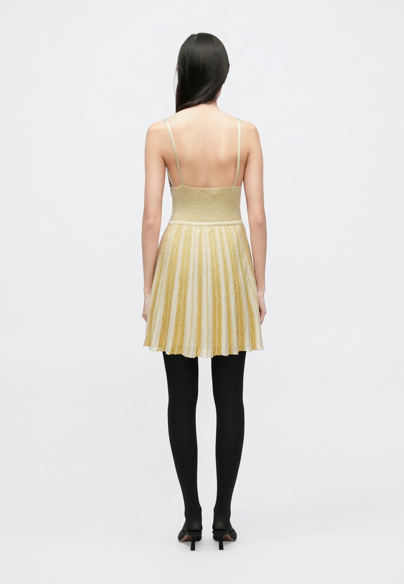 Strickkleid mit einem eng anliegenden goldenen Oberteil und einem ausgestellten, plissierten Rock. Der Rock zeigt abwechselnd goldene und weiße Streifen. Wird mit schwarzen Strumpfhosen getragen.