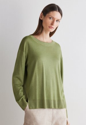 Kvinde med glat brunt hår iført løs lysegrøn sweater og beige bukser, stående mod en ensfarvet hvid væg med hænderne i lommerne.