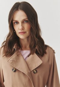 Beige Trenchcoat mit doppelter Knopfleiste, großen Knöpfen und großem Kragen. Weicher Stoff mit dezenter Textur.