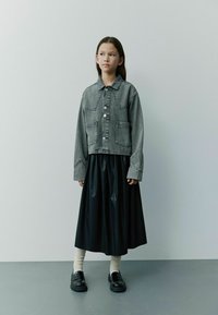 Harmaa farkkutakki suurilla taskuilla, musta keinonahkahame, vaaleat ribbisaappaat ja mustat loafersit, neutraalin taustan edessä.