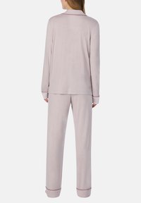 Vrouw in een lichtroze lange mouwen top en losse broek in pyjamastijl, staand met de rug naar de camera tegen een effen achtergrond.