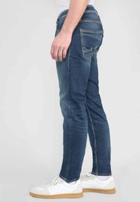 Jeans in denim blu con una vestibilità slim, tasche standard e cuciture gialle, abbinati a sneakers bianche con suole in gomma e dettagli colorati.