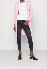 Cárdigan largo rosa claro, camiseta blanca, jeans ajustados oscuros y mules negros con tacones cuadrados; textura suave, diseño ajustado y longitud corta.