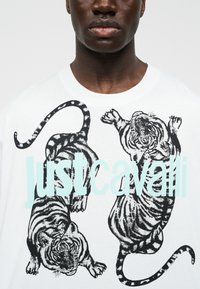Άνδρας που φοράει λευκό T-shirt με μαύρα σχέδια τίγρης και ανοιχτό μπλε κείμενο "Just Cavalli" στο στήθος σε απλό φόντο.