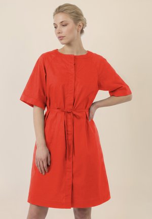 Frau mit blondem Haar in einem niedrigen Dutt, die ein knielanges, knallrotes Kleid mit kurzen Ärmeln und einem gebundenen Taillengürtel trägt, steht vor einem beigen Hintergrund.