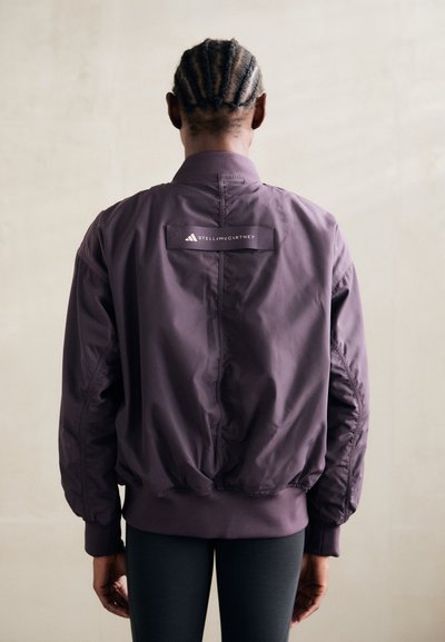 Lila bomber dzseki, testhez simuló aljjal, hátsó emblémával és sima, könnyű anyaggal. Rugalmas mandzsettákkal és oldalsó varrással rendelkezik.