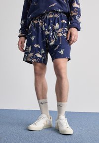 Mørkeblå svømmebukser med et farverigt flamingo-mønster, elastisk talje og sidelommer, kombineret med hvide sneakers og cremefarvede sokker.