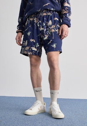 Navy swim shorts featuring a colorful flamingo print, elastic waistband, and side pockets, paired with white sneakers and cream socks.