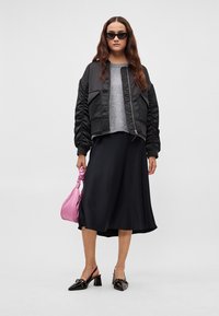 Sort bomberjakke, grå sweater, sort nederdel, sorte lak slingback-hæle og en lyserød håndtaske. Model med langt bølget hår og solbriller.