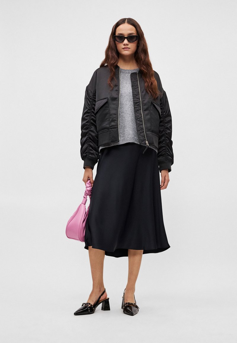 Sort bomberjakke, grå sweater, sort nederdel, sorte lak slingback-hæle og en lyserød håndtaske. Model med langt bølget hår og solbriller.