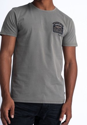 Mand iført en grå T-shirt med korte ærmer og et lille mørkt grafisk mærke på venstre bryst, kombineret med mørke bukser.