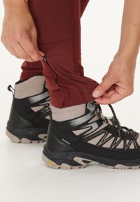 Schwarze wasserdichte Wanderschuhe mit grauen Akzenten; bordeauxfarbene Hosen mit seitlichen Reißverschlüssen; Hände, die die Hosenabschlüsse über die Schuhe anpassen.