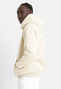 Sudadera beige con un diseño amplio, que cuenta con un gran bolsillo frontal, una capucha ajustable con cordón y texto sutilmente grabado en la parte de atrás.