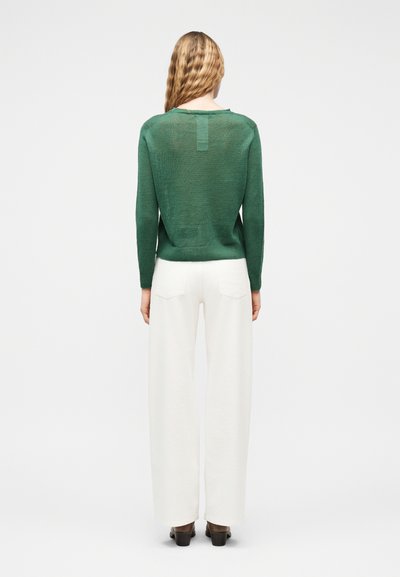 Femme aux cheveux blonds ondulés portant un pull vert à manches longues, un pantalon blanc à larges jambes et des chaussures foncées, debout de dos sur fond blanc.