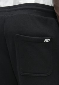 Pantaloni de trening negri cu un buzunar pe spatele pantalonilor, având un mic logo alb oval aproape de marginea superioară.