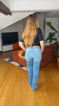 Γυναίκα με μακριά ξανθά μαλλιά, που φοράει μαύρο πουκάμισο και μπλε τζιν, στέκεται σε σαλόνι με ξύλινο πάτωμα και καφέ καναπέ.