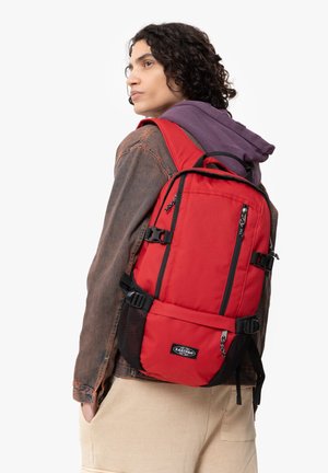 Mochila de tela roja con acentos negros, correas ajustables y bolsillos laterales de malla. Presenta múltiples cremalleras y un asa en la parte superior.