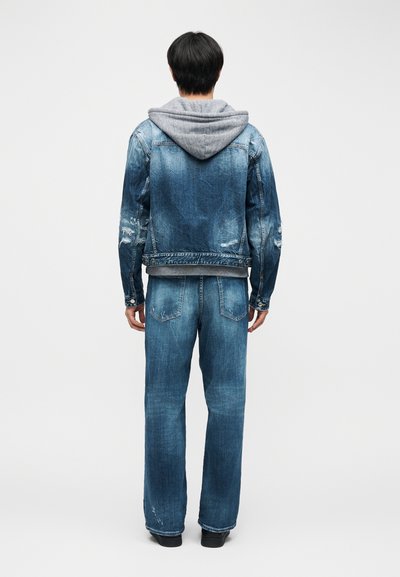 Άνδρας που στέκεται γυρισμένος με την πλάτη, φορώντας ξεθωριασμένο μπλε τζιν μπουφάν πάνω από γκρι φούτερ με κουκούλα και τζιν παντελόνι σε ίδιο ύφασμα, με μαύρα παπούτσια, σε απλό φόντο.