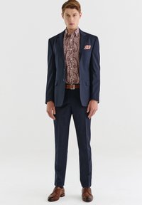 Granatowy garnitur z klapami w kształcie litery V, sparowany z koszulą w wzór paisley w odcieniach czerwieni i kremu, brązowy skórzany pasek i pasujące buty.