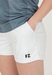 Vita atletiska shorts med en slät yta, med en svart 'FZ'-logotyp på den nedre vänstra sidan. I kombination med en grå topp med prickmönster.