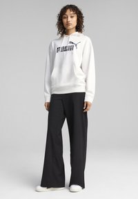 Hoodie bianco con logo PUMA nero, abbinato a pantaloni neri a gamba larga e sneakers bianche. Tessuto morbido e design dalla vestibilità rilassata.