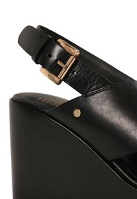 Schwarze Lederkeilsandale mit einem goldenen Schnallenriemen und kleinem rundem goldenen Logodetail an der Seite.
