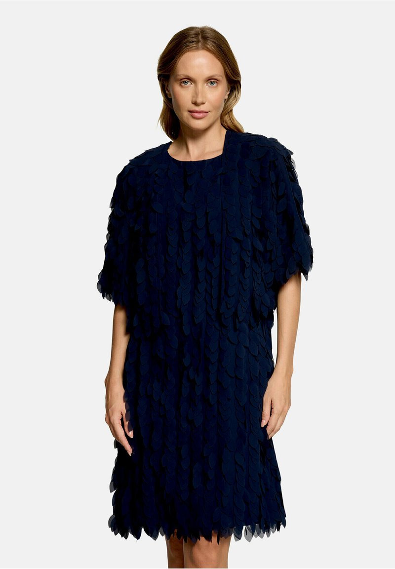 Femme aux cheveux clairs portant une robe bleu foncé jusqu’aux genoux, recouverte de pièces de tissu superposées en forme de pétales, debout devant un fond uni.