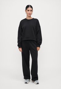 Donna con capelli neri raccolti, che indossa una felpa nera, pantaloni larghi neri, sneakers bianche e orecchini a cerchio argentati, in piedi su uno sfondo bianco.